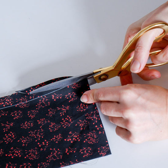 Hands cutting a piece of black fabric with red patterns using gold scissors on a white background