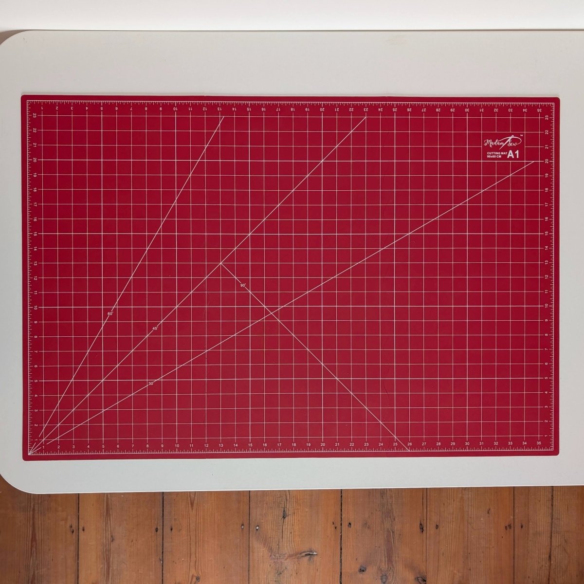 A red cutting mat with white grid lines and diagonal guide lines, displayed on a white surface over a wooden floor.