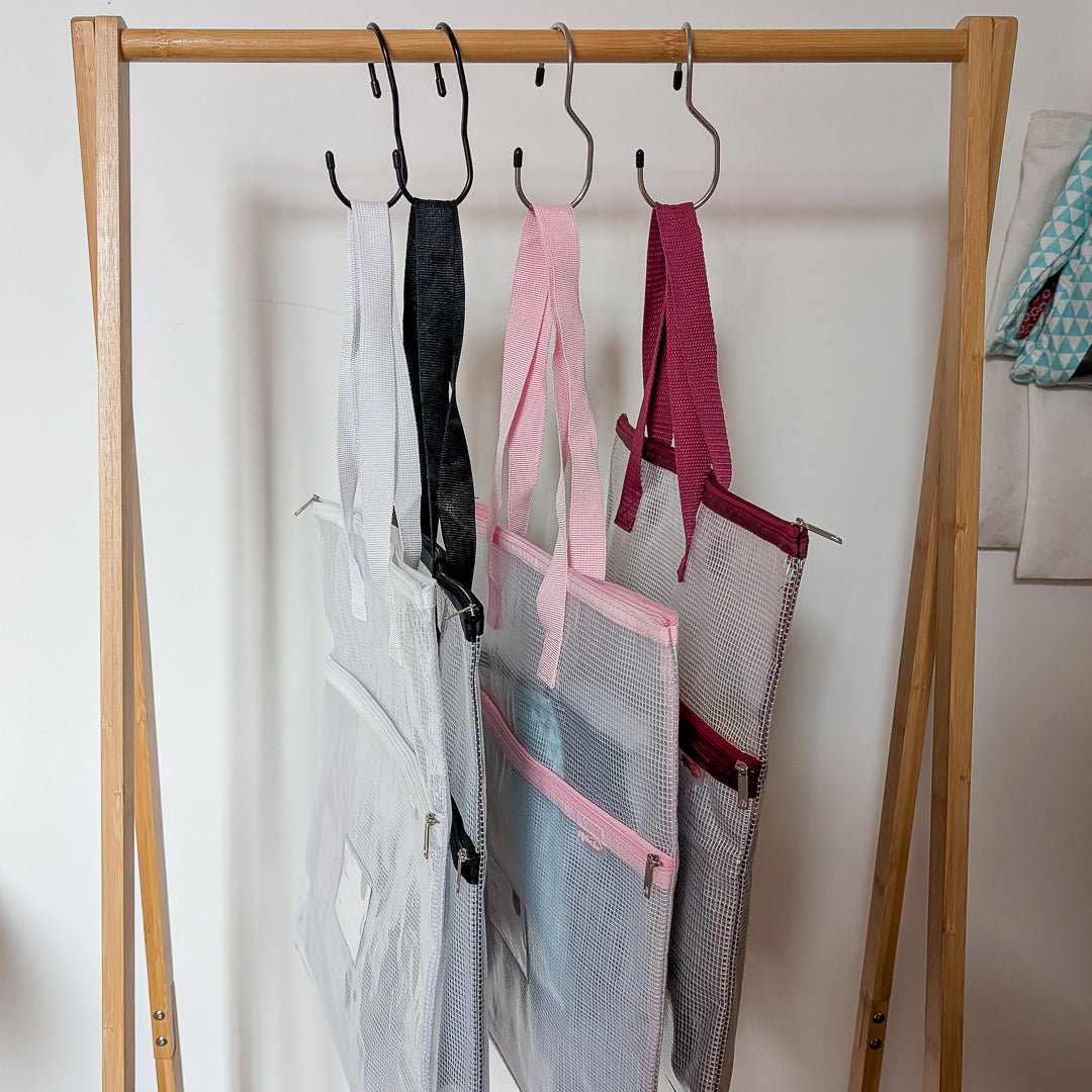White, black, pink and burgundy mesh project bags hanging from hooks on a wooden rail