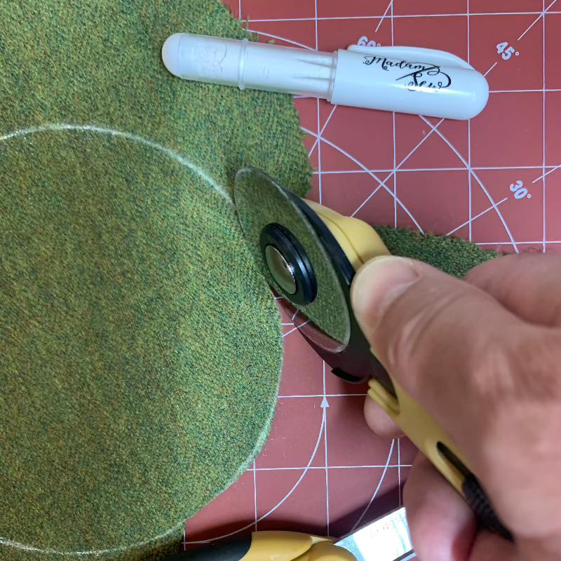 Cutting a circle out of green fabric with a rotary cutter folliwing a white mark drawn with a chalk marking pen