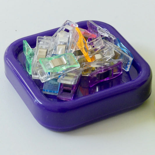 A pile of small translucent plastic clips in assorted colors, including purple, green, blue, orange, and clear, arranged inside a square purple dish on a white background.