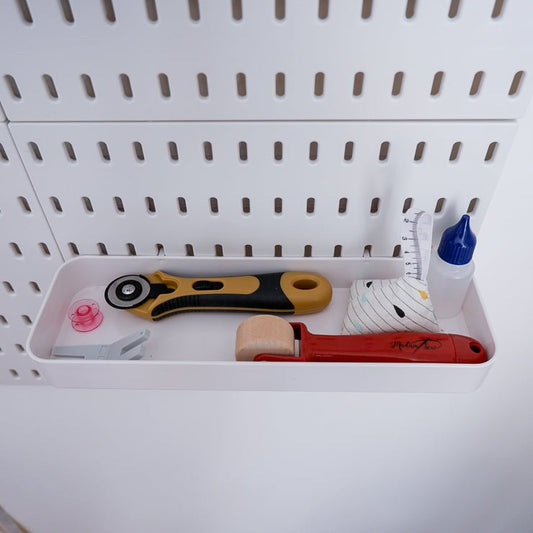 White long plastic tray attached to a peg board holding a rotary cutter, a seam roller, some glue, a pattern weight