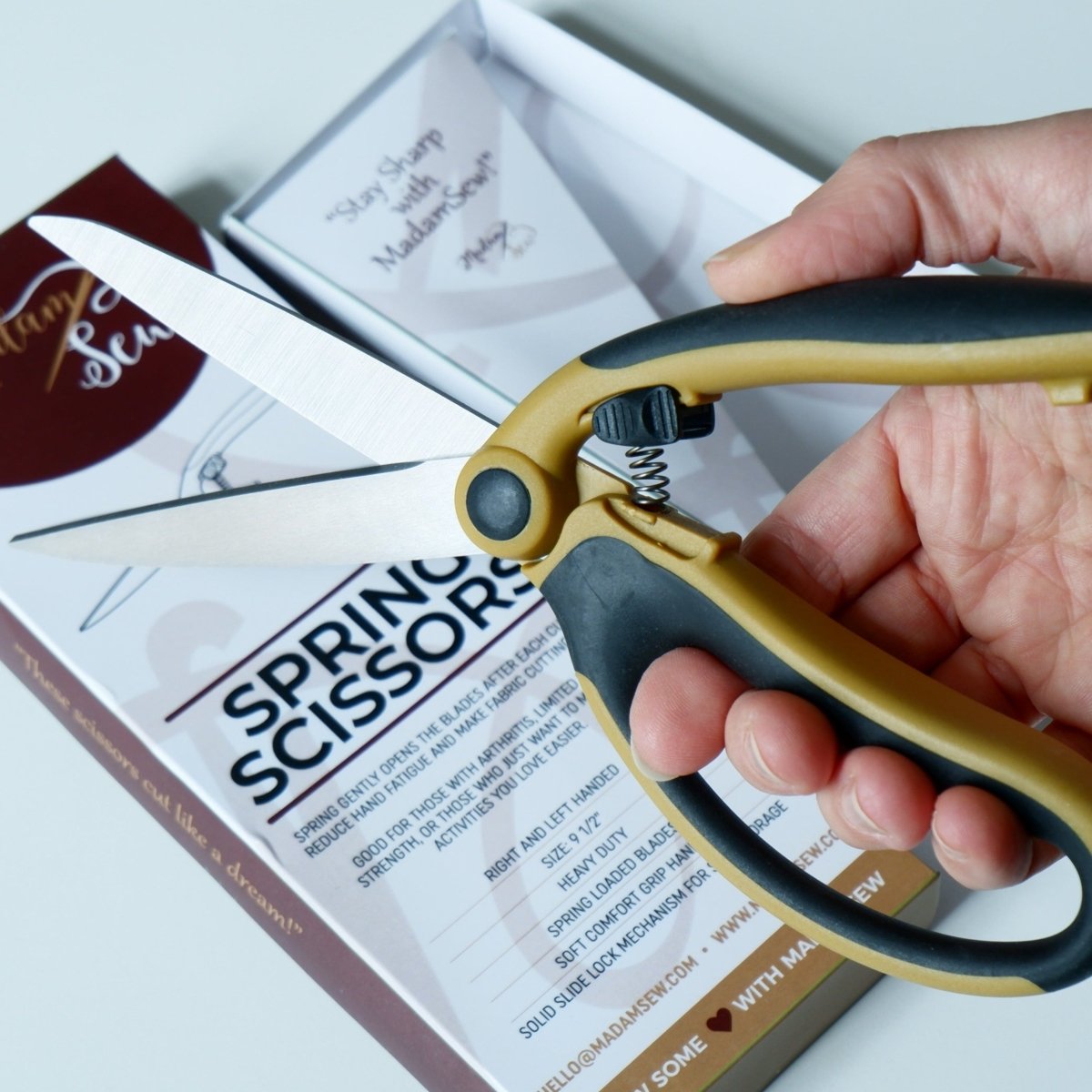 A hand showing the Madam Sew spring scissors in the open position with the packaging in the back